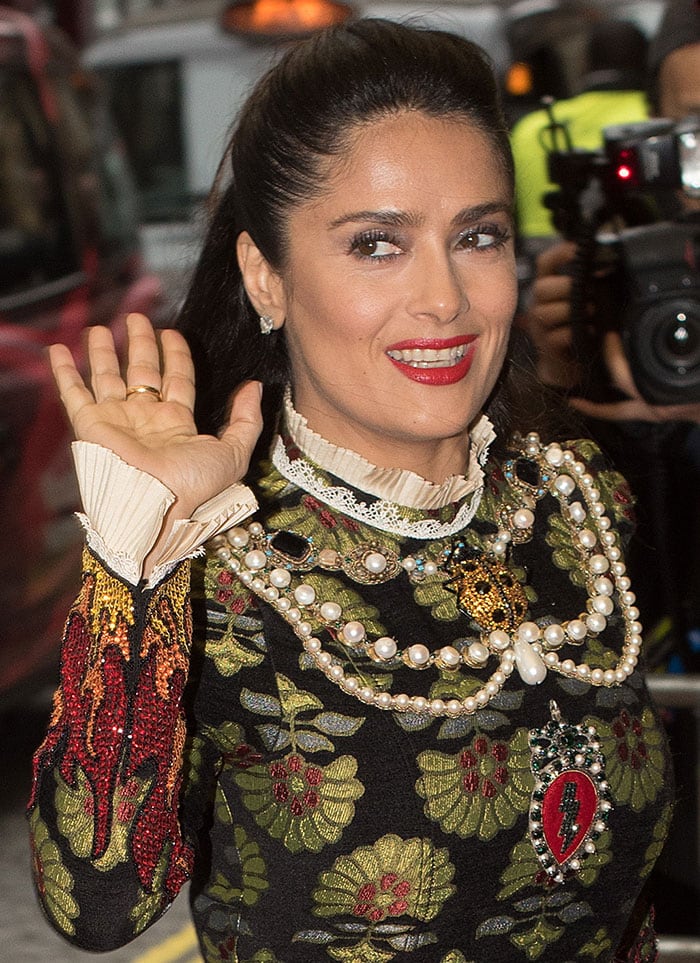 Hayek waves to photographers, highlighting her flawless glam and the ornate jeweled accents across her neckline