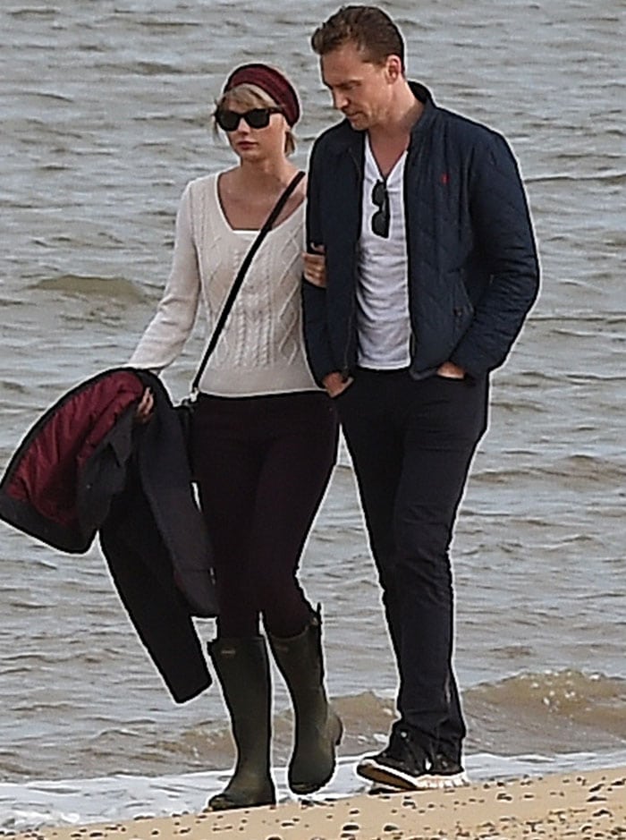 Taylor keeps it understated yet polished in Le Chameau “Chasseur” boots, Black Orchid Malaga Wine jeggings, a cream cable-knit sweater, a Free People “Myna” headband, Ray-Ban Wayfarers, and a Rebecca Minkoff “Mara” saddle bag during a seaside stroll with Tom Hiddleston