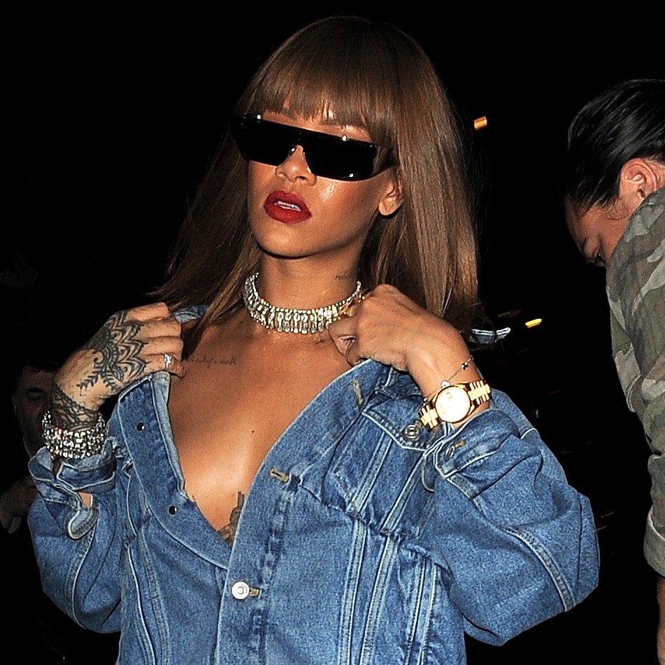 Rihanna styled her oversized denim jacket off the shoulders for a daring nightlife look, pairing the plunging silhouette with futuristic Dior sunglasses and layered diamond jewelry outside Tape Nightclub in London