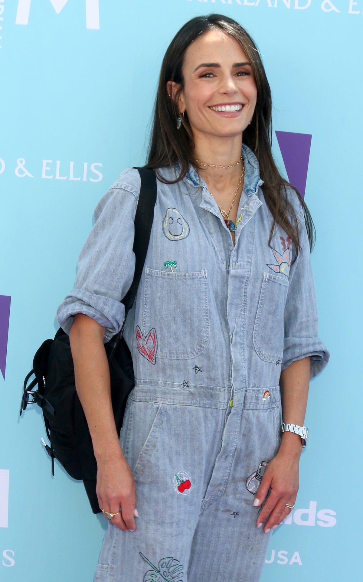 Jordana Brewster keeps it playful in a light denim jumpsuit decorated with whimsical embroidered patches, paired with layered necklaces and a casual black backpack