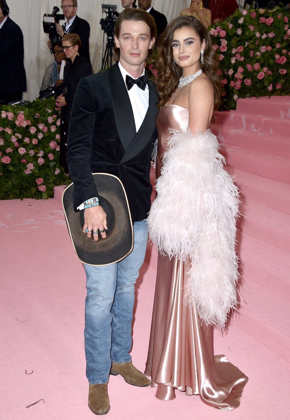 Standing 5'9" (175.3 cm) beside 6'½" (184.2 cm) Patrick Schwarzenegger, Taylor Hill brought old-Hollywood glamour to the 2019 Met Gala in a blush satin Ralph Lauren gown styled with diamond jewelry and a dramatic feather boa, while Schwarzenegger added a modern Western twist in cowboy boots, jeans, and a tuxedo jacket topped with a black hat