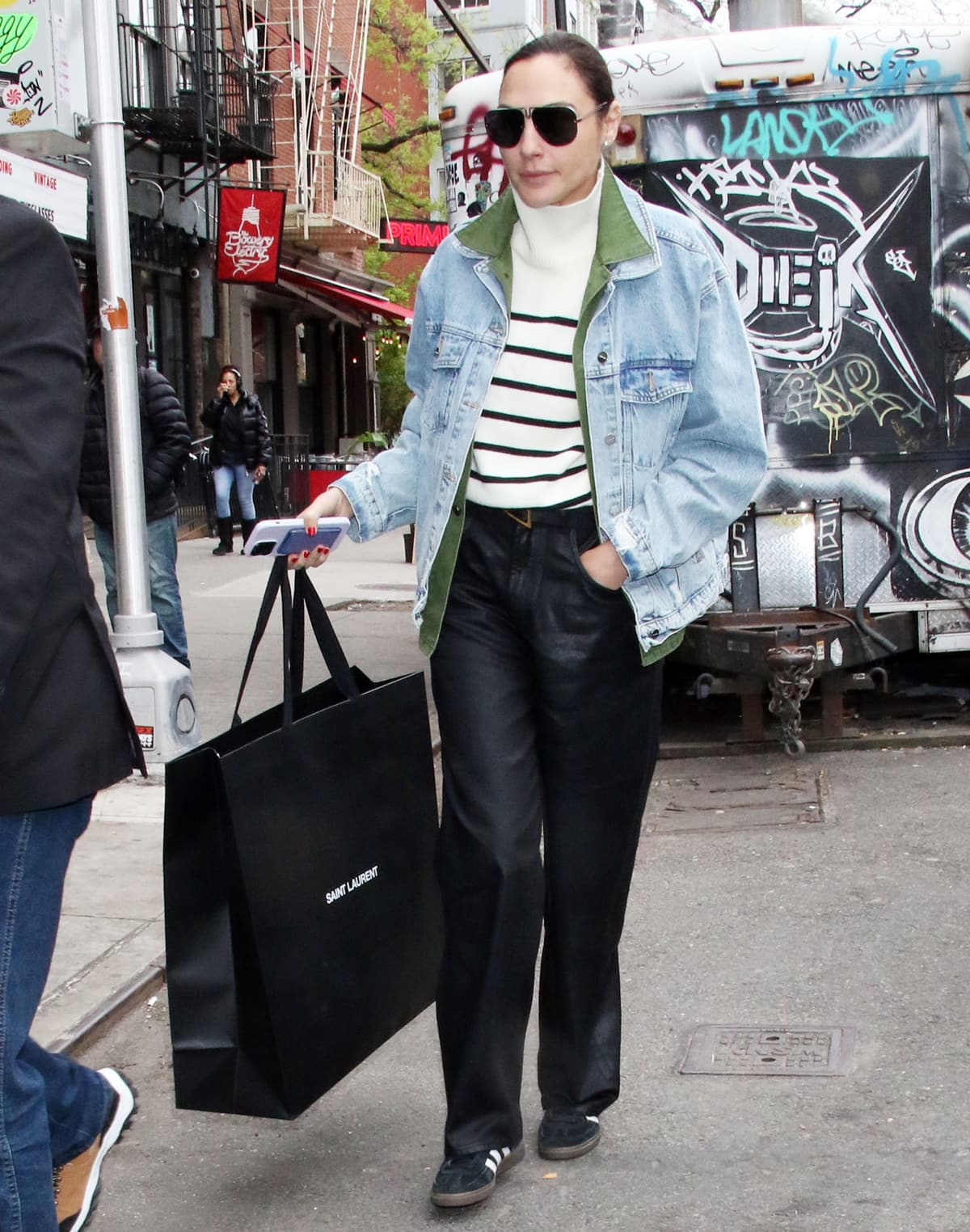On April 28, 2023, in New York City, Gal Gadot stepped out in a chic layered look—pairing a black-and-white turtleneck sweater, khaki overshirt, and faded denim jacket with relaxed leather trousers and classic black adidas sneakers