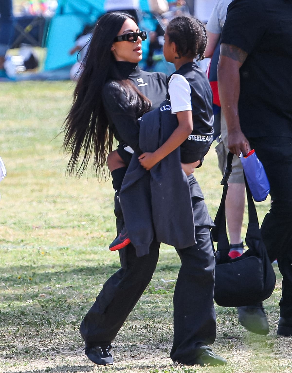 Kim Kardashian carries her son Saint West while wearing a Balenciaga sporty B bike jersey top, LED sunglasses, and Yeezy Boost 700 “Vanta” sneakers during a soccer outing in Los Angeles
