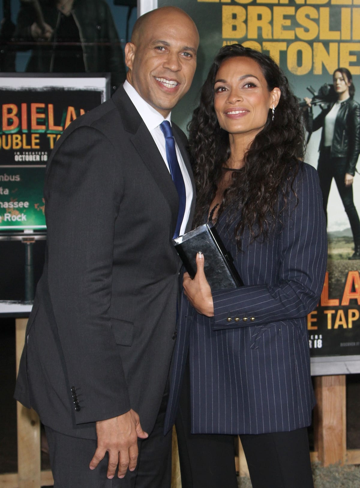 Rosario Dawson leans in beside Cory Booker at the Zombieland: Double Tap premiere, pairing her pinstripe blazer and sleek black trousers with a glossy clutch and natural, wavy hair for a polished, modern finish