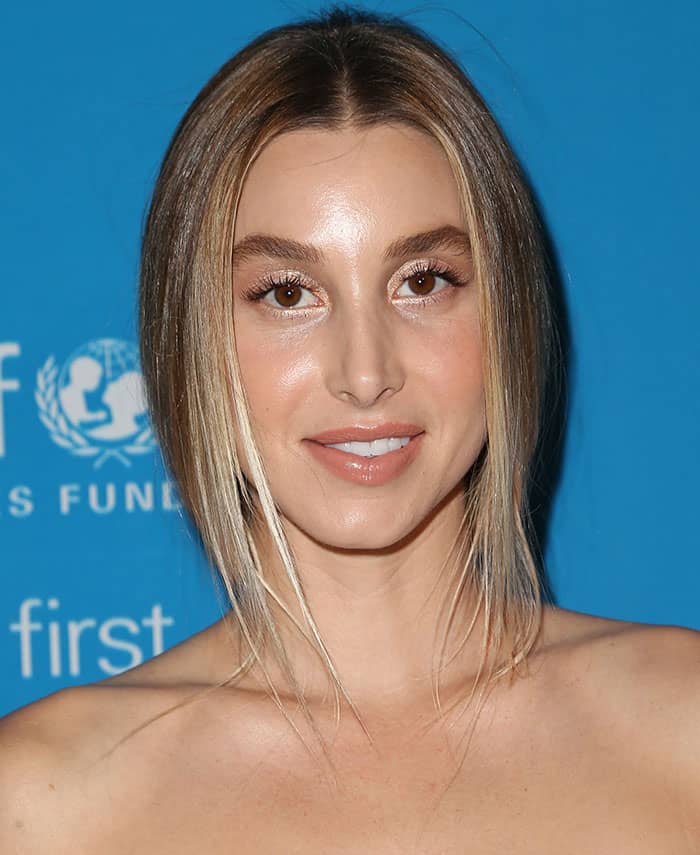 Whitney Port completes her look with sleek, center-parted hair and a radiant makeup palette featuring metallic eyeshadow and a soft peach lip