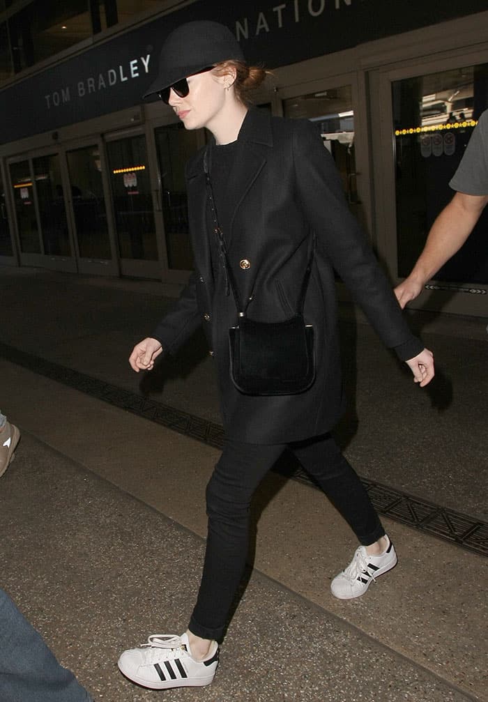 Emma Stone keeps it effortlessly chic at LAX, pairing a classic black peacoat and skinny jeans with white Adidas 'Superstar' sneakers, completing her look with a riding cap and Ray-Ban sunglasses for an understated, cool vibe