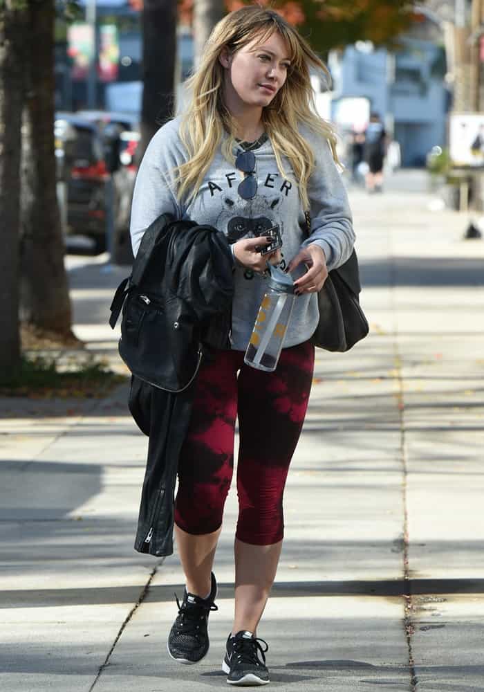 Hilary Duff keeps it sporty and stylish in red tie-dye capri leggings paired with a cozy raccoon sweatshirt, black Nike sneakers, and a leather jacket in hand