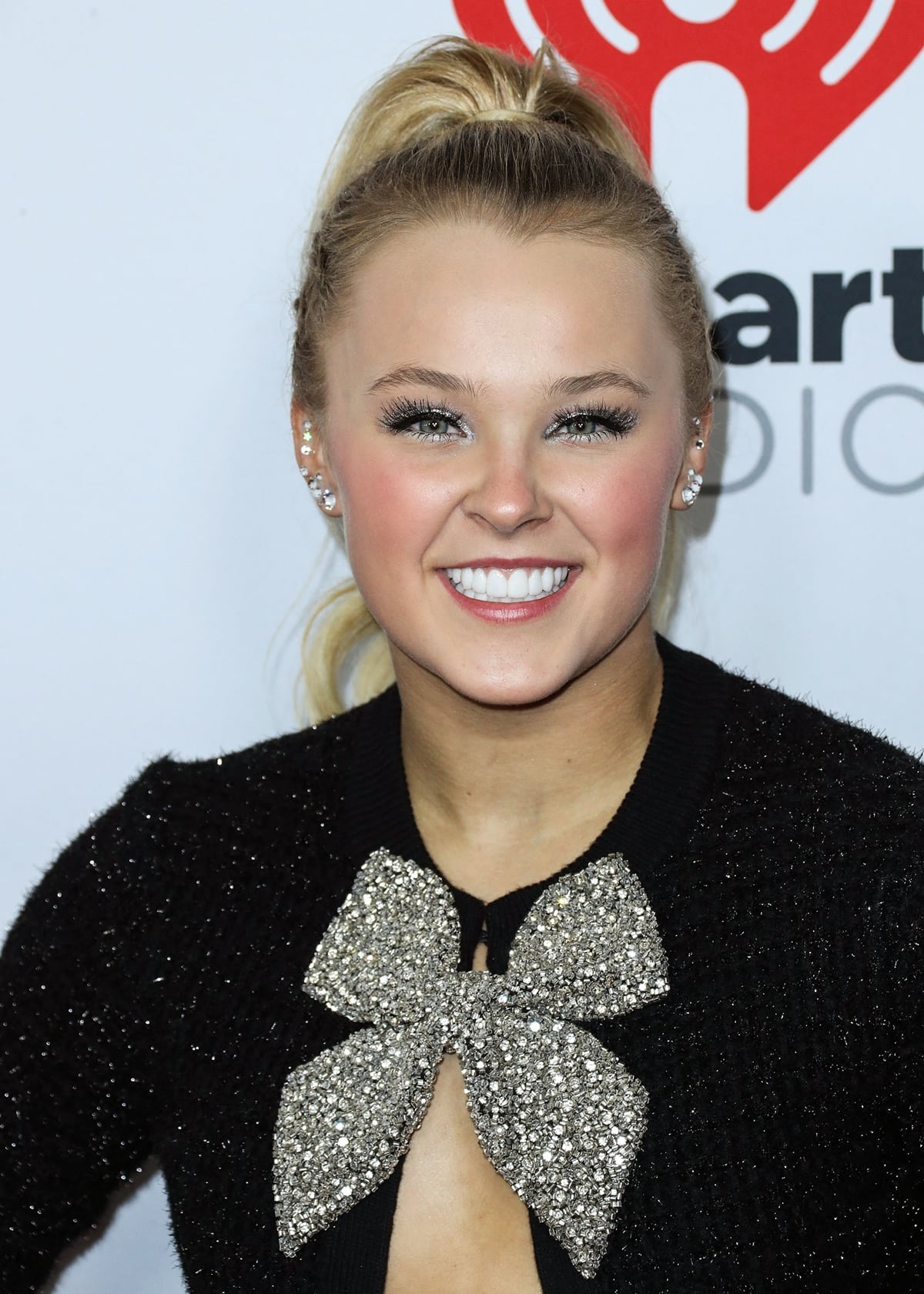 JoJo Siwa smiles on the red carpet with a sleek high ponytail, shimmering eye makeup, and a statement crystal bow detail at her neckline