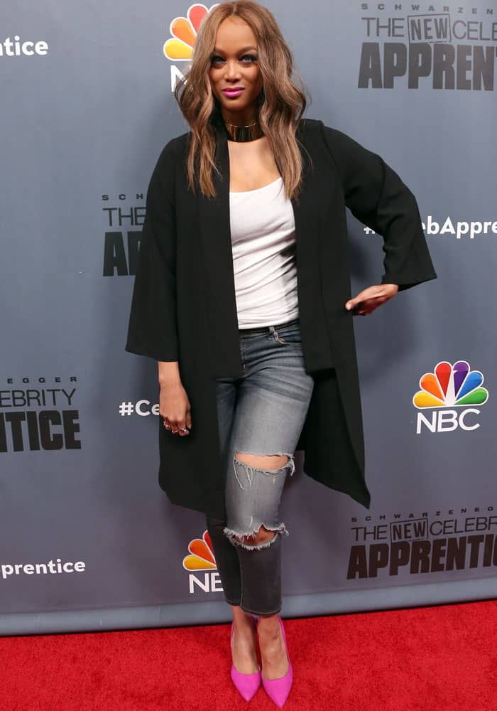 Tyra Banks poses on the red carpet for "The New Celebrity Apprentice" wearing a casual yet chic outfit with ripped jeans, a long black blazer, and vibrant pink heels