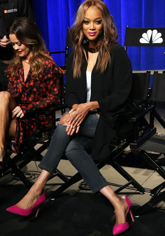 Tyra Banks sits with her legs crossed, wearing pink heels and a black blazer during a panel event for The New Celebrity Apprentice