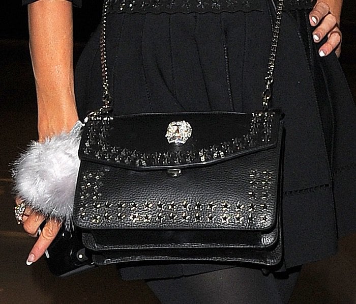 Paris Hilton accessorizes her outfit with a luxe black leather shoulder bag adorned with studded detailing, a chain strap, and a silver embellishment for a touch of edgy elegance