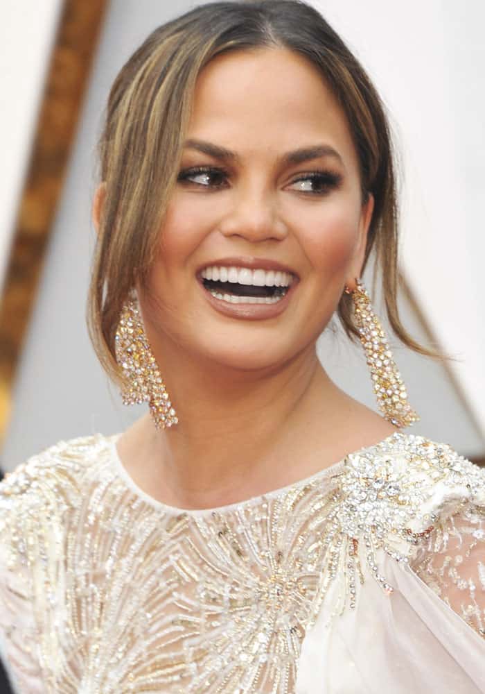 Chrissy Teigen stuns with her radiant smile, sleek updo, and dazzling chandelier earrings that perfectly complement her intricately beaded gown and smoky eye makeup