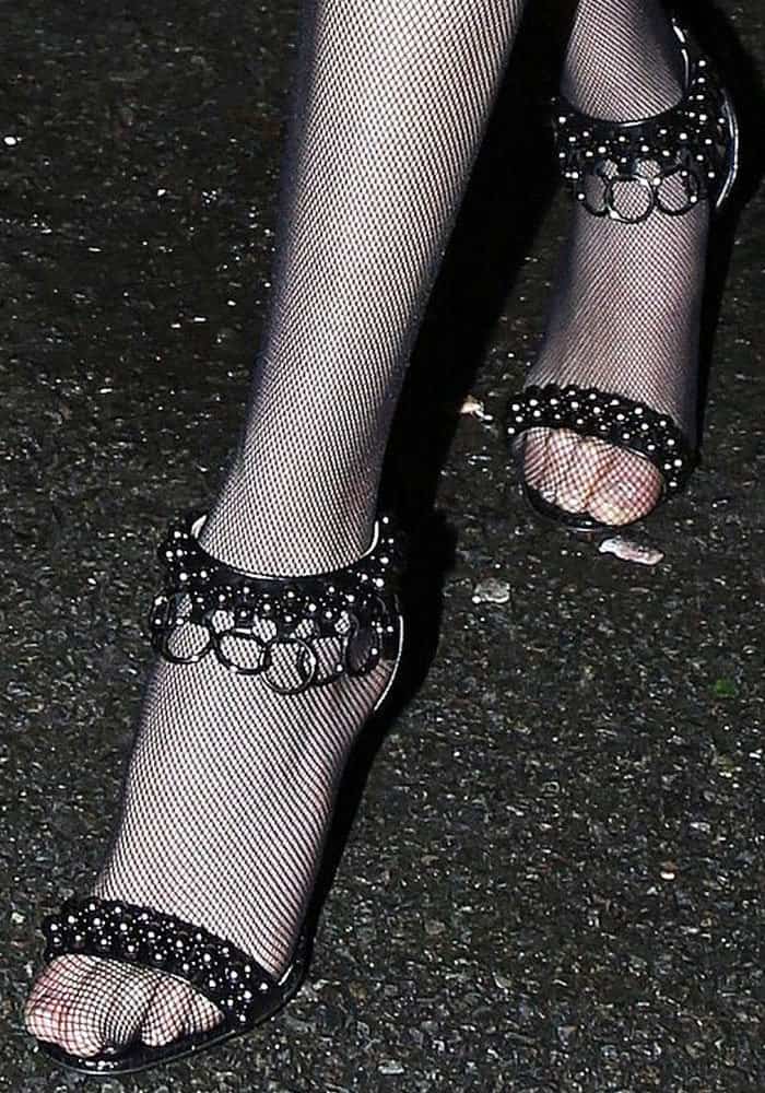 Bella Hadid completes her edgy Parisian look with black Christian Louboutin Gypsandal heels, featuring studded embellishments and chain-link details, effortlessly styled with fishnet stockings for a bold, high-fashion finish