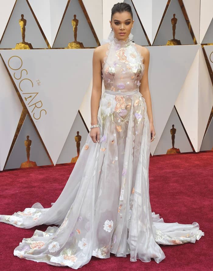 To replicate Hailee Steinfeld's Oscars look, opt for a sheer halter gown with floral appliqués, delicate ruffles, and soft, minimal accessories for a timeless red carpet vibe