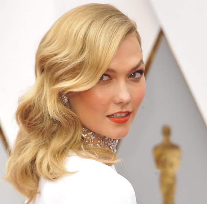 Karlie Kloss exudes old-Hollywood glamour with her soft side-swept waves, bold orange lipstick, and a dazzling statement choker that elevates her Oscars look