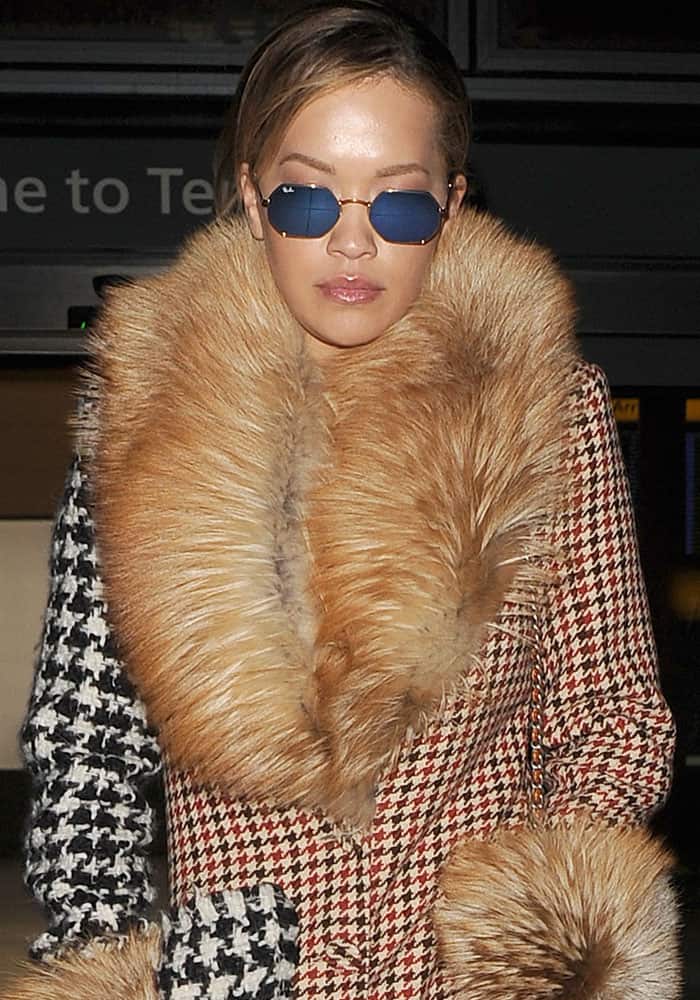 Rita Ora complements her eclectic ensemble with classic Ray-Ban Hexagonal sunglasses and a lavish fur-trimmed Ellery coat, emphasizing her fearless approach to airport fashion