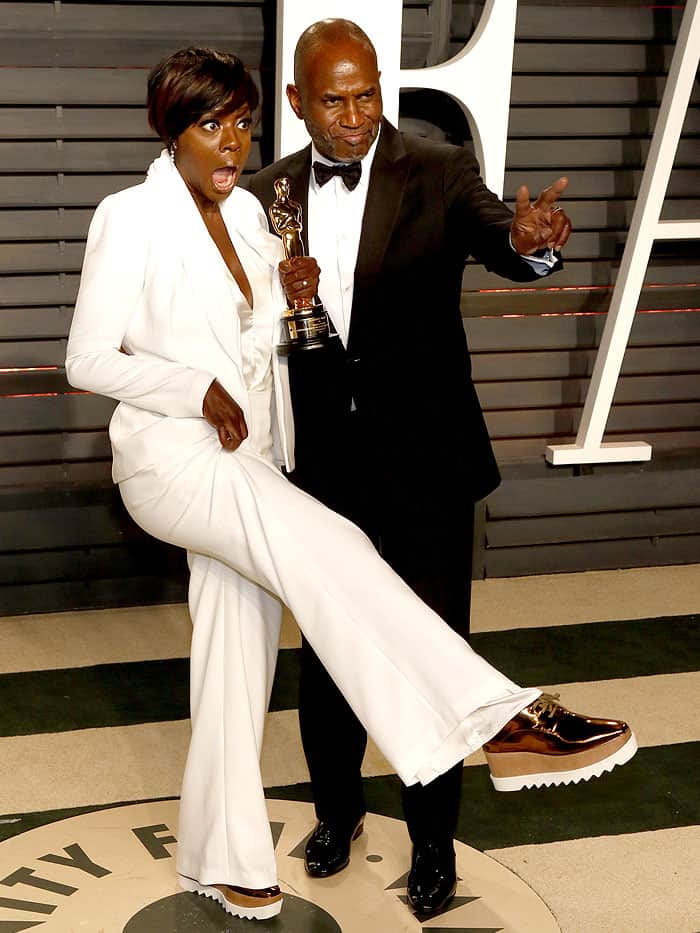 Viola Davis stuns in a crisp white tailored pantsuit paired with gold Stella McCartney ‘Elyse’ platform oxfords, while her husband, Julius Tennon, keeps it classic in a sharp black tuxedo with a bow tie, creating the perfect red carpet power couple look