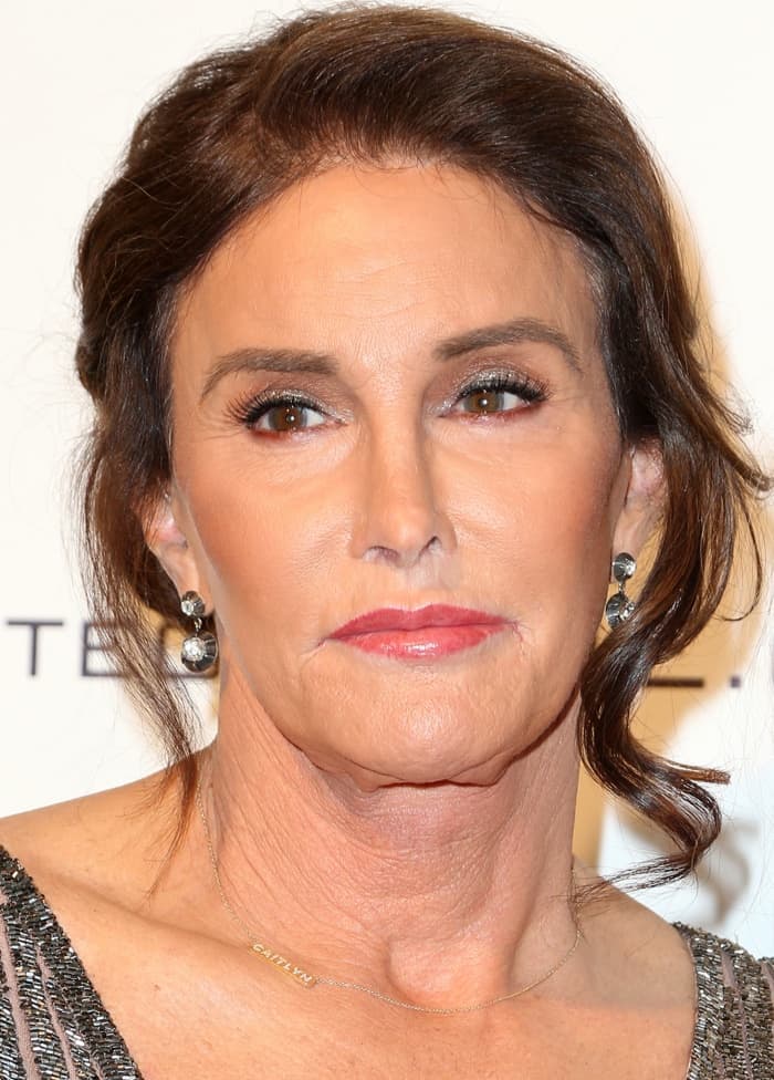 Caitlyn Jenner stuns with a chic side-swept updo and soft tendrils, wearing elegant drop earrings from Ronda Kamihira paired with a delicate gold nameplate necklace