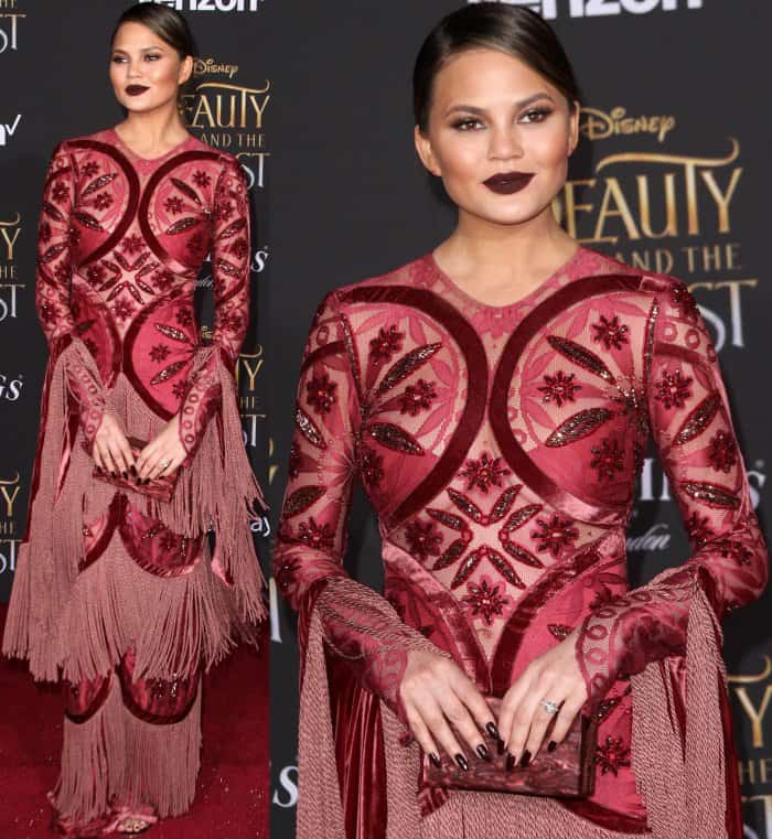 Chrissy Teigen completes her vampy Raisa Vanessa look with a sleek side-parted ponytail, a matching dark red Edie Parker clutch, and statement earrings from EF Collection
