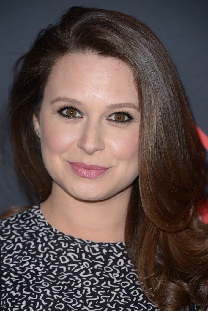 Katie Lowes' rich chocolate brown hair is styled in long, voluminous waves, complementing her light brown eyes
