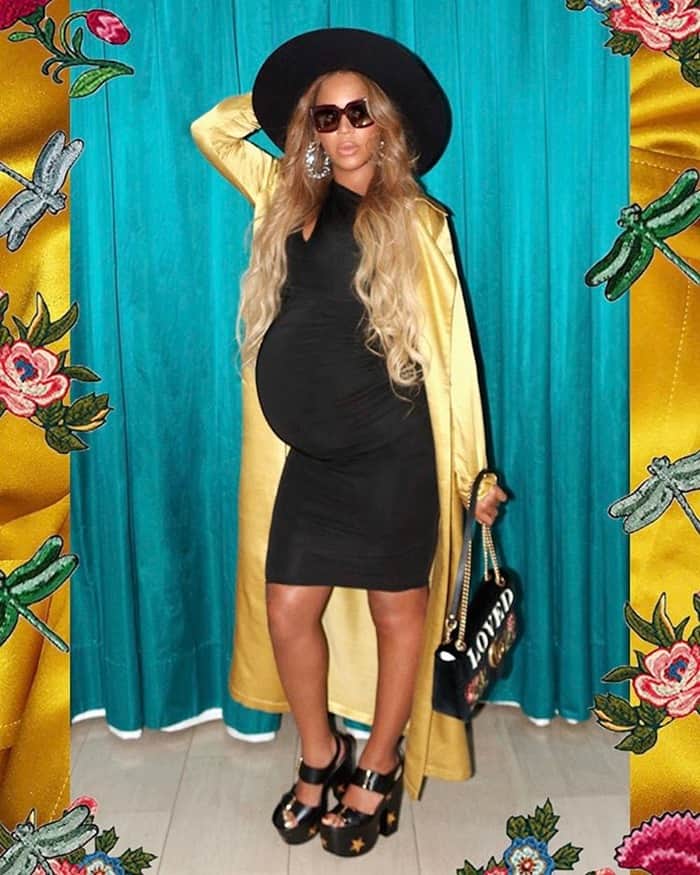 Beyoncé poses confidently in a sleek black maternity dress layered with a gold trench coat, grounding the look with star-studded platform sandals that add height and attitude