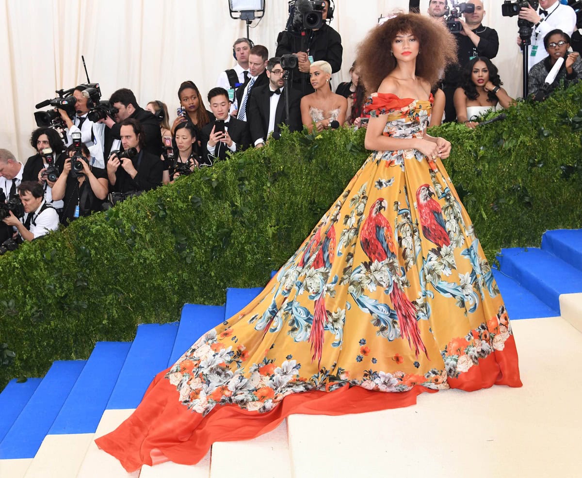 Zendaya stuns at the 2017 Met Gala in a vibrant off-the-shoulder Dolce Gabbana Alta Moda gown adorned with tropical parrots and a sweeping orange-lined train