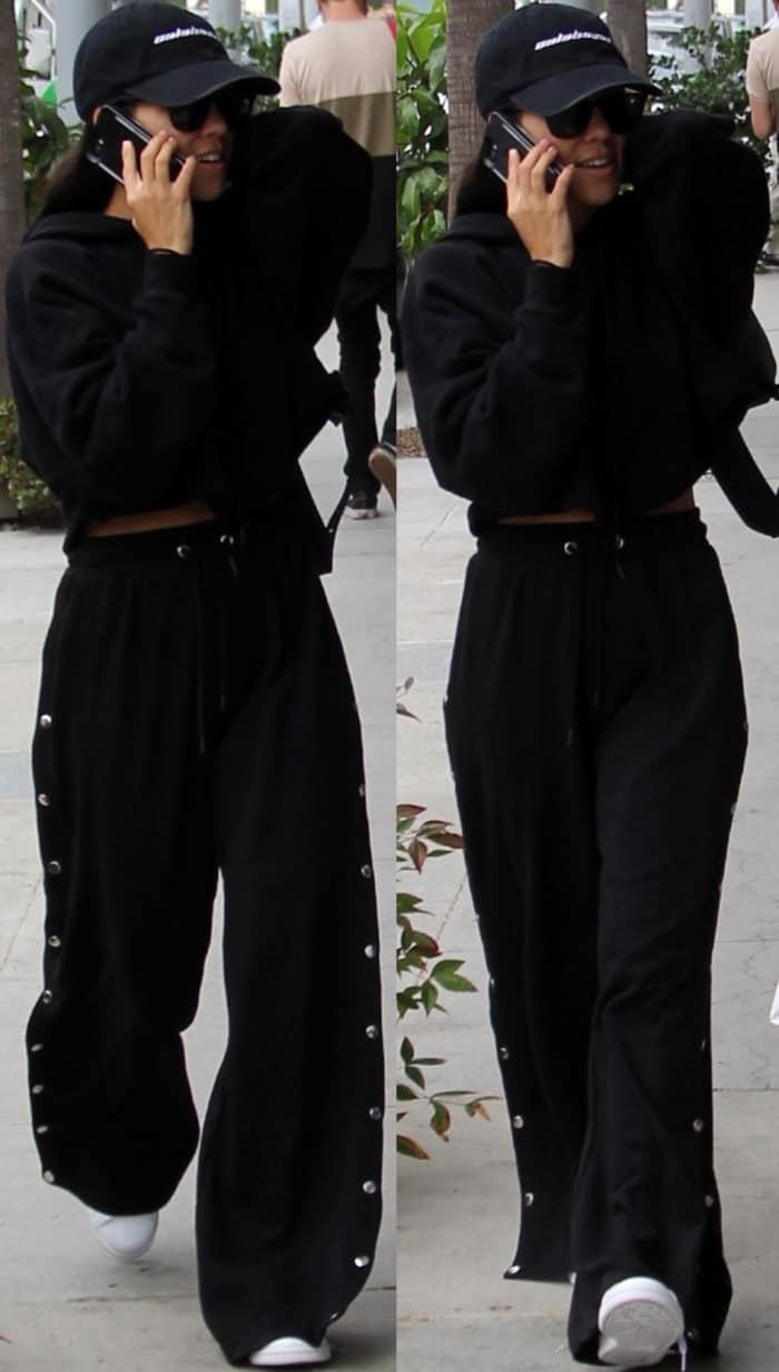 Kourtney Kardashian keeps it casual in a cropped black hoodie, snap-side track pants, a baseball cap, and classic white sneakers while out in Beverly Hills