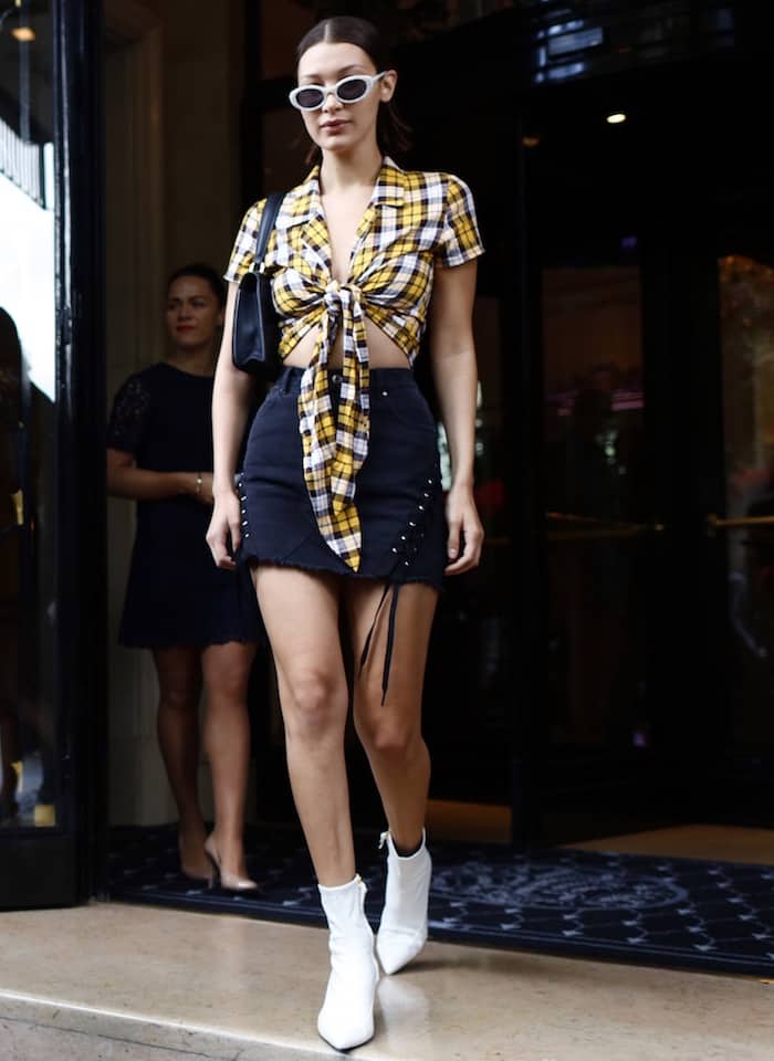 Bella Hadid exits a building in a tied plaid crop top, a dark denim mini skirt with lace-up details, white ankle boots, and cat-eye sunglasses