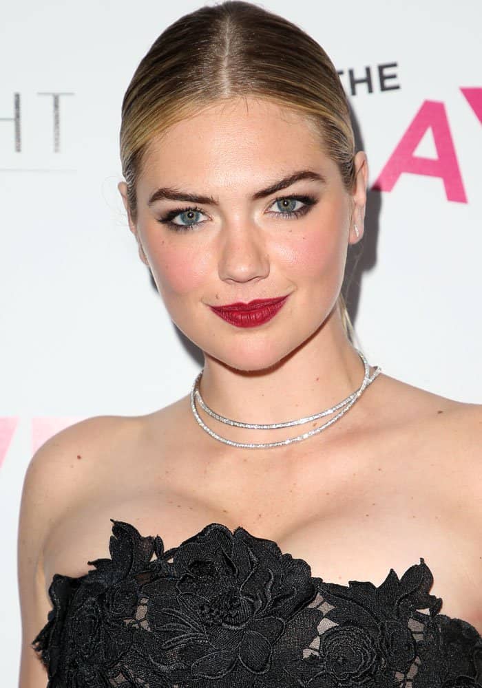 Kate Upton radiates elegance with a sleek center-parted low bun, bold brows, smoky eye makeup, and deep red lips, complementing her sparkling layered diamond choker and strapless black lace J. Mendel gown