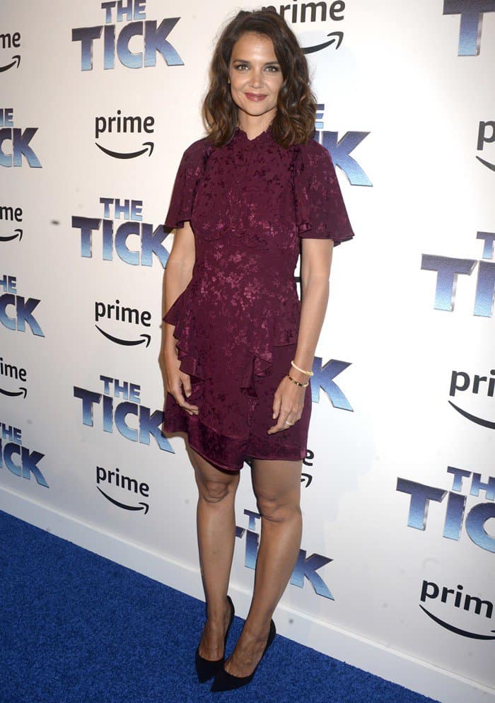 Posed against the branded backdrop, Holmes’ burgundy mini dress stands out for its layered detailing and refined fit, delivering quiet elegance on the New York premiere circuit