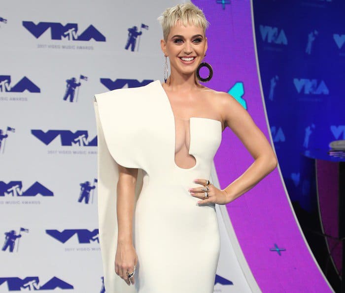 Katy Perry turns heads in a sculptural white Stéphane Rolland couture gown featuring a bold sternum cutout, asymmetrical cape sleeve, and oversized hoop earrings