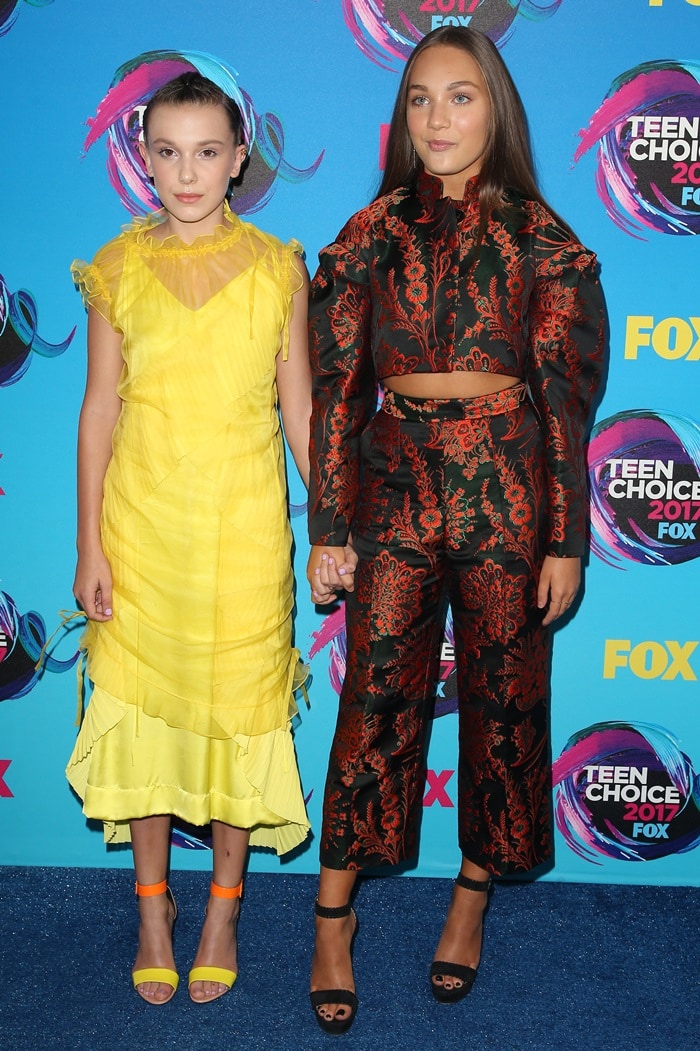 Millie Bobby Brown and Maddie Ziegler hold hands on the blue carpet at the 2017 Teen Choice Awards at USC’s Galen Center in Los Angeles on August 13, 2017, their contrasting silhouettes and heel heights subtly highlighting the difference in proportion between Millie’s bright, ankle-strap sandals and Maddie’s sleek black heels