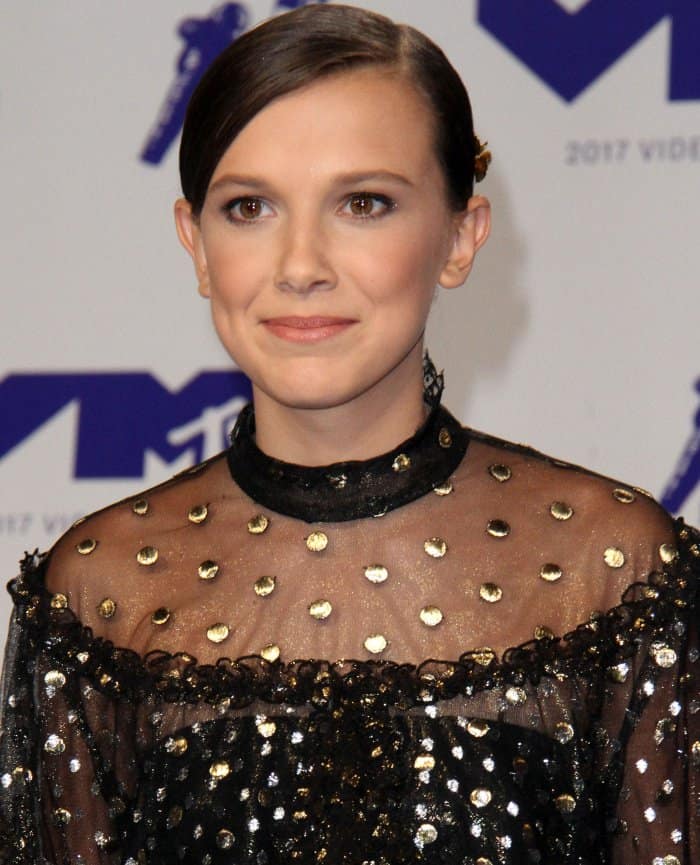 A sleek side-swept updo and subtle makeup highlight Millie’s youthful take on the edgy, gold-studded Rodarte neckline