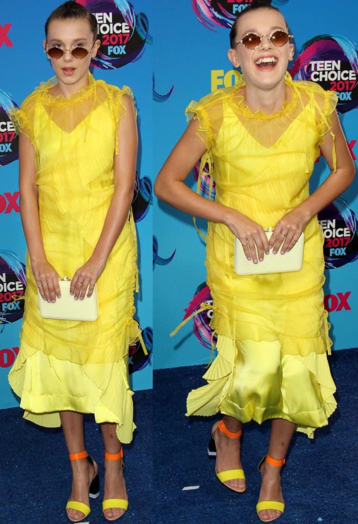 Millie Bobby Brown poses front-on in her ruffled yellow Kenzo dress, clutch in hand, letting the sheer layers and bright palette set the tone against the blue carpet