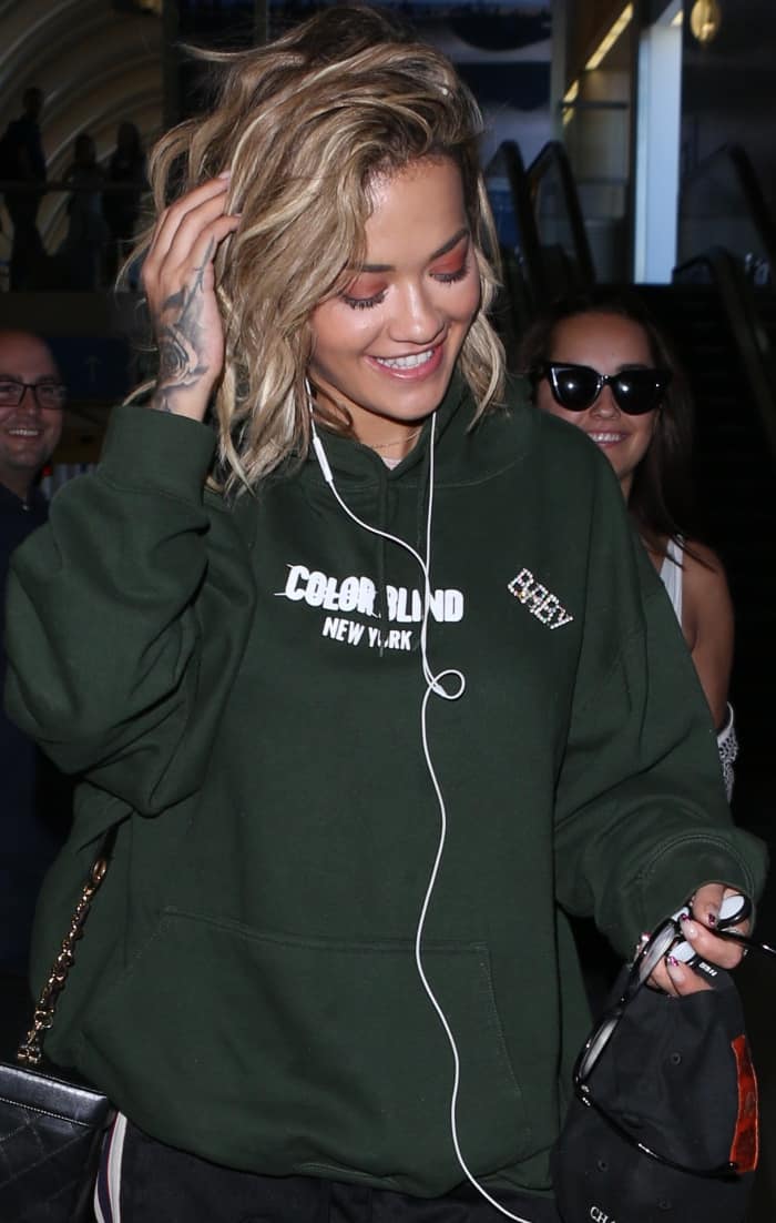 Rita Ora wearing a dark green hoodie, black trackpants, and white Reebok "Classic Leather" sneakers at LAX