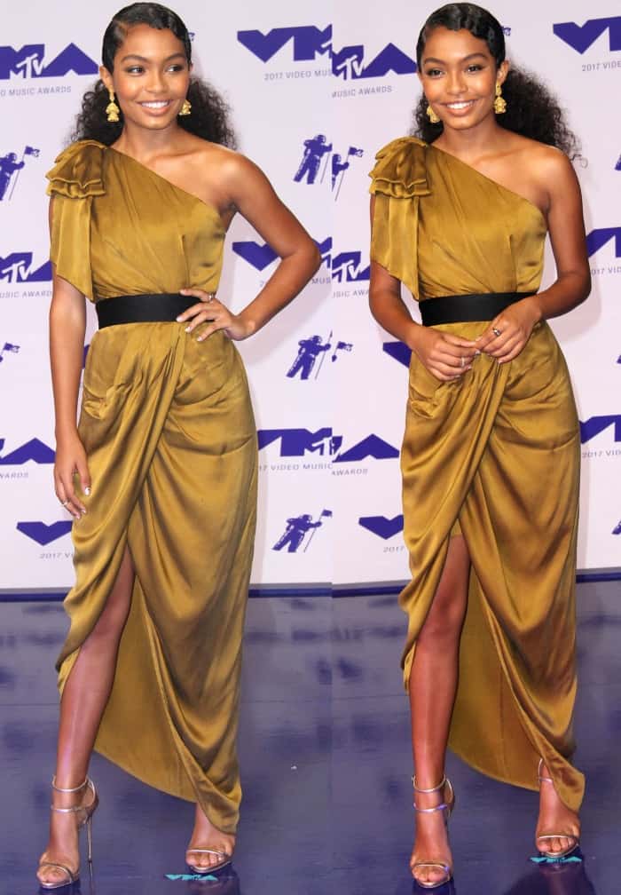 Yara Shahidi exudes elegance in a one-shoulder, draped gold Zimmermann gown, complemented perfectly by metallic Giuseppe Zanotti 'Darcie' sandals and statement earrings