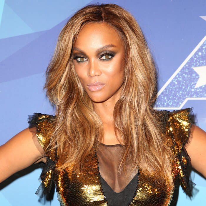 Tyra Banks pairs bronzed glam makeup with loose waves, complementing the high-shine texture of her gold sequined mini dress