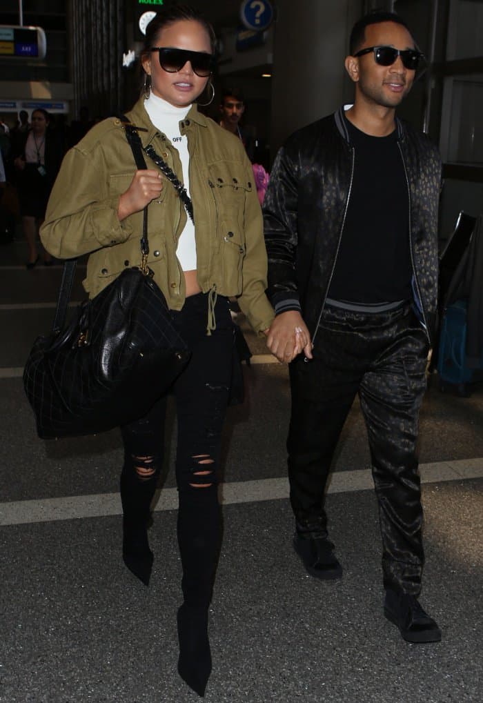 Chrissy Teigen and John Legend walk hand-in-hand through LAX, with Chrissy showcasing her cropped jacket and Balenciaga sock boots alongside John’s patterned tracksuit