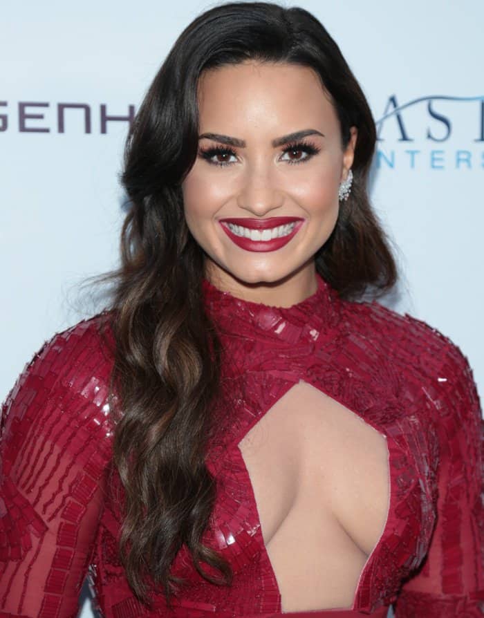 A close-up on Demi Lovato's stunning beauty look, featuring her glamorous classic waves, a bold red lip to match her gown, and sparkling diamond stud earrings