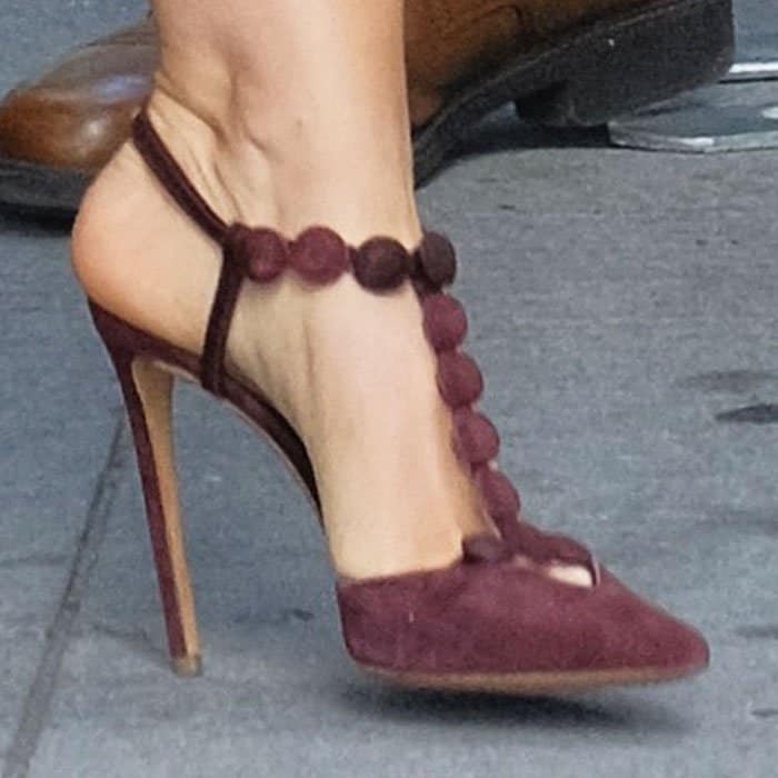 Isla Fisher's feet in purple suede Nicholas Kirkwood pumps, featuring a playful T-strap design adorned with circular pom-pom embellishments for a bold yet whimsical touch