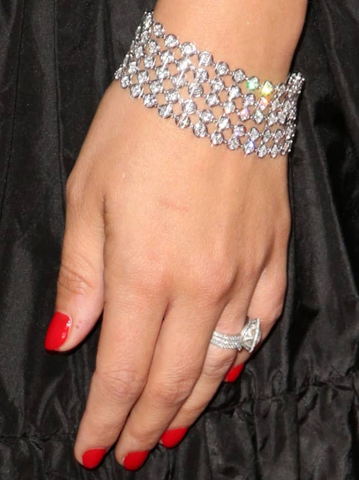 Natalie Portman adds high-shine contrast with Van Cleef Arpels diamond jewelry, layering a sparkling lattice bracelet with a multi-band ring against the dark Dior fabric and glossy red manicure