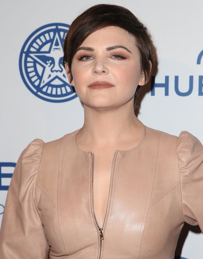 A close-up of Goodwin shows her soft glam makeup and cropped hairstyle complementing the sleek neckline of the leather dress