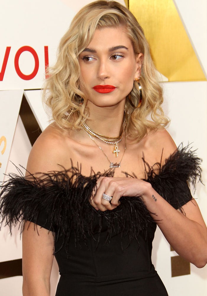Hailey Baldwin accessorizes her off-shoulder feathered mini dress with layered gold necklaces, including a cross pendant and her nameplate, as she confidently adjusts her outfit on the red carpet
