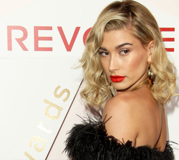 Hailey Baldwin exudes glamour as she showcases the back of her feathered mini dress, complemented by oversized gold hoop earrings and a bold red lip