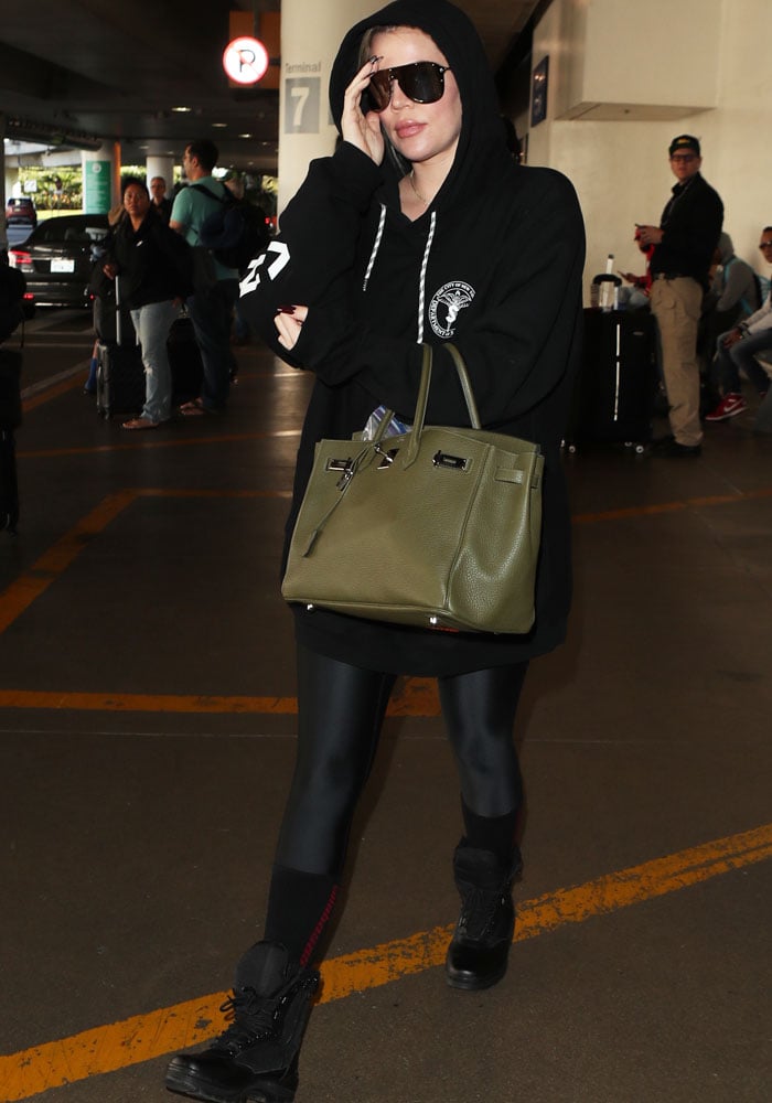 Khloé Kardashian navigates the airport in a signature blend of comfort and luxury, featuring an oversized Heron Preston hoodie, sleek leggings, combat boots, and an Hermès Birkin bag