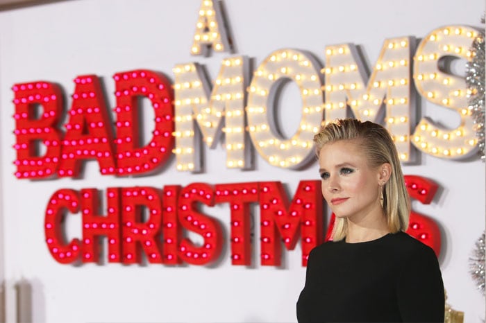 Kristen Bell, with her sleek hairstyle and elegant attire, poses gracefully beside the vibrant, light-up promotional signage for her film, "A Bad Moms Christmas"