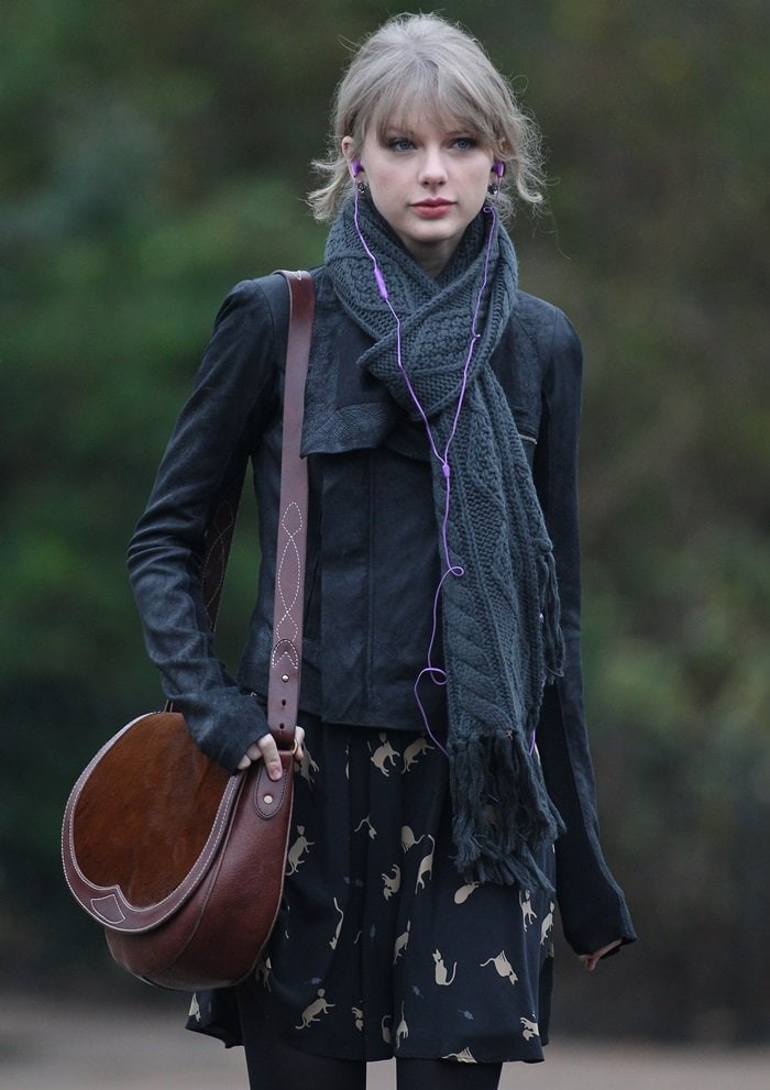 Taylor Swift elevates her London street style with a classic brown Ralph Lauren saddle bag, perfectly complementing her edgy black suede jacket, cozy knit scarf, and whimsical cat-print dress