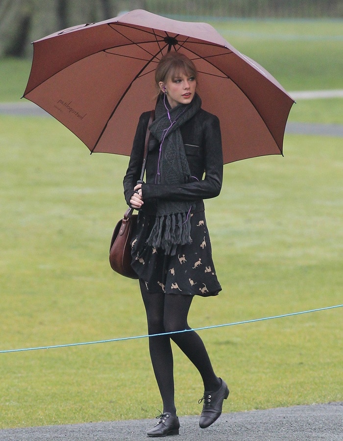 Taylor Swift braves the London rain in chic black patent leather Chanel oxfords, pairing them effortlessly with black tights, a playful cat-print dress, and a cozy scarf for a polished yet relaxed look