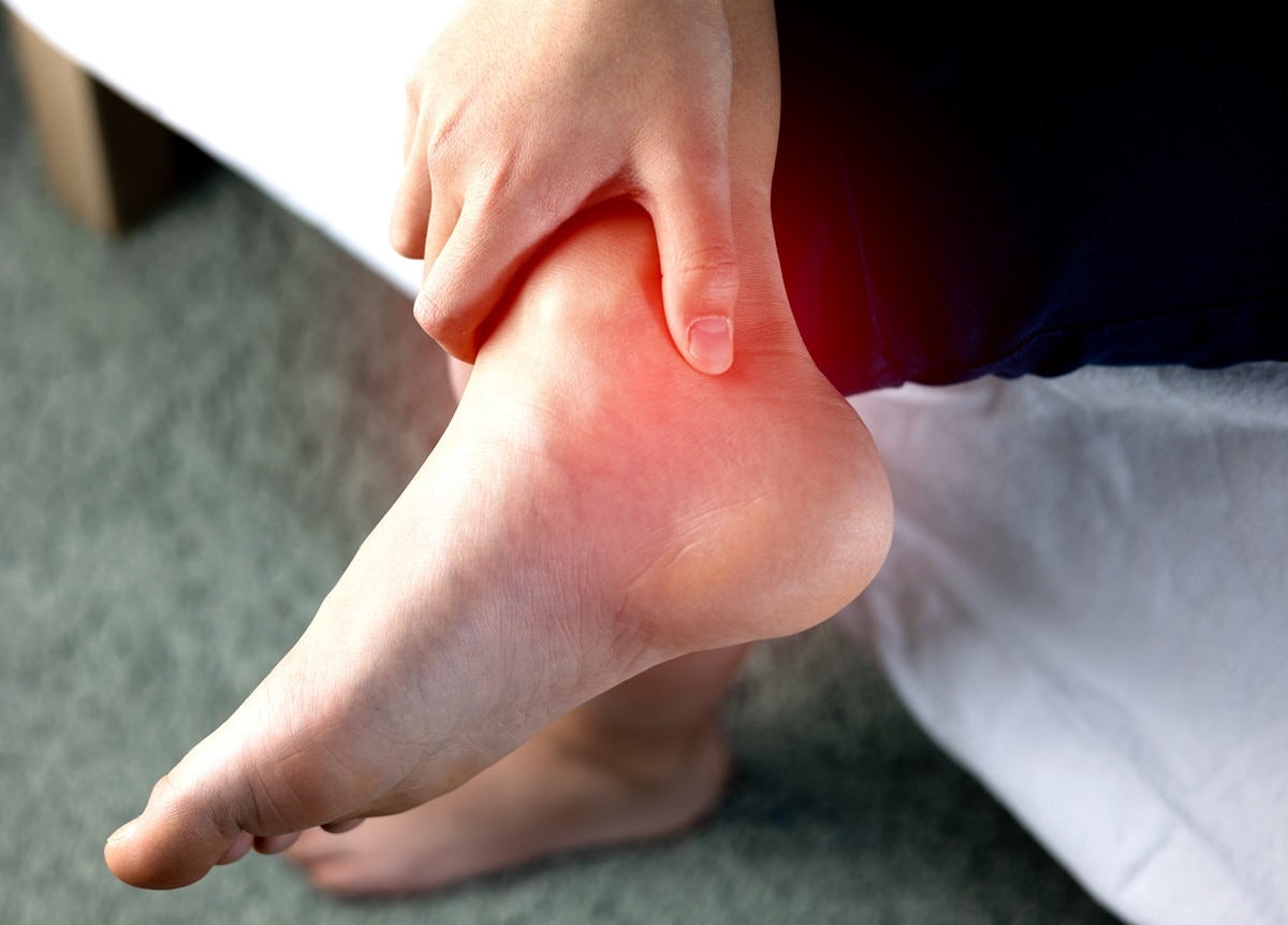 A person gripping their ankle near the Achilles tendon, highlighting discomfort and pain in the inflamed red area, possibly indicating tendonitis or strain