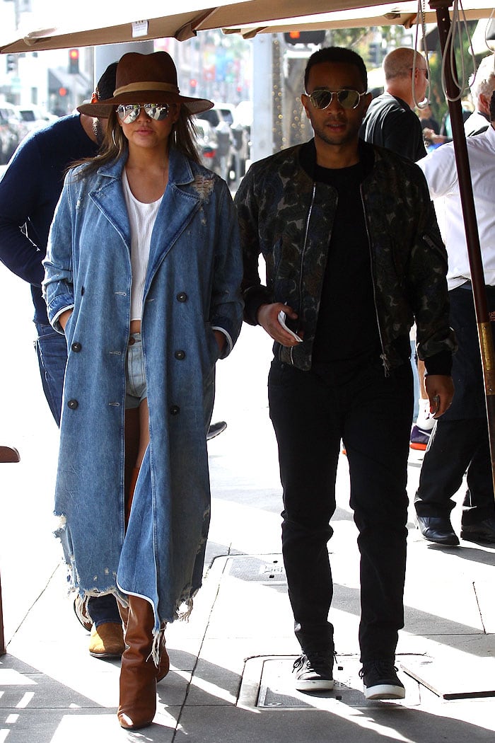 Chrissy Teigen steps out with John Legend in Beverly Hills, flashing her baby bump in a loose white cropped tank and denim short shorts, styled with a distressed Alexander Wang trench coat and Saint Laurent “Niki” thigh-high boots